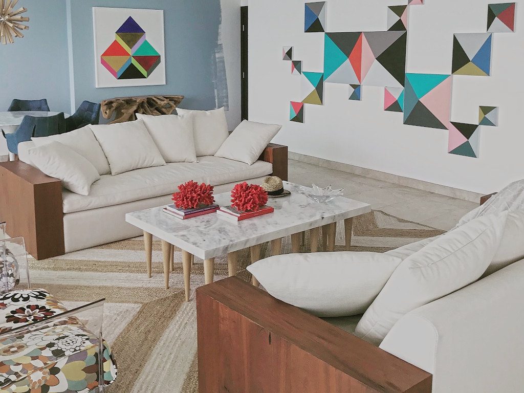 Living room with white sofas in red-wood frames, a marble coffee table with red coral, a wall of geometric works and a blue painted wall