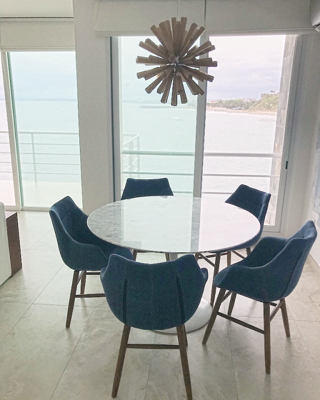 A round marble table with blue chairs under a wooden sunburst pendant, the beach through the window