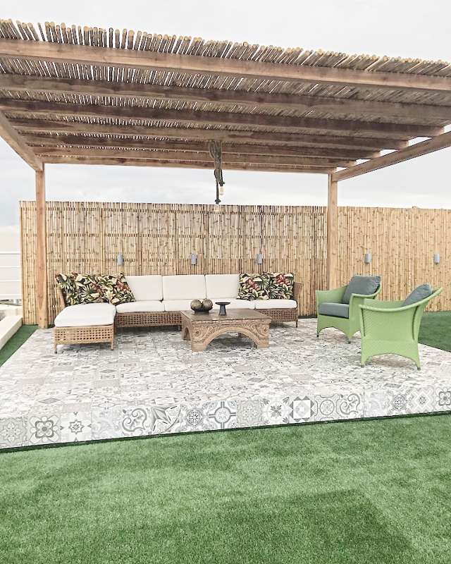 Under a cane pergola, a wicker sectional with white cushions, green armchairs and a carved table on patterned tiles