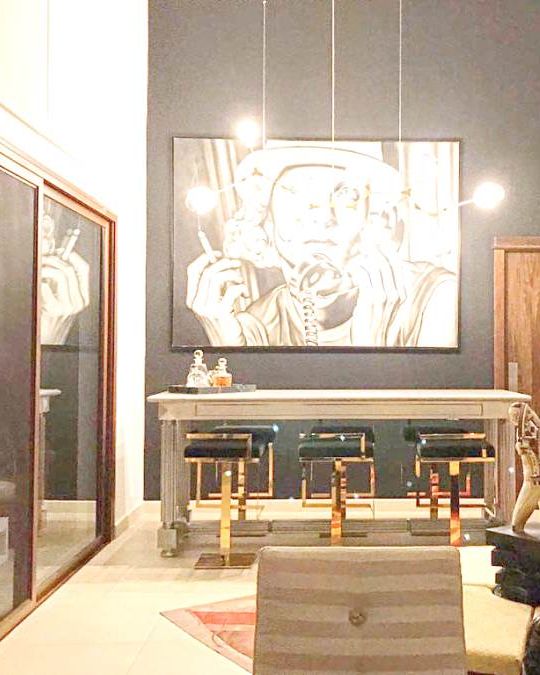 A marble bar with gold and black stools against a black wall, under a large black and white portrait and globe lights