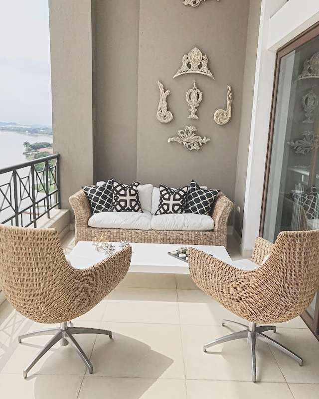 A terrace with a wicker sofa, two wicker swivel chairs and carved wood ornaments on a taupe wall, the river beyond