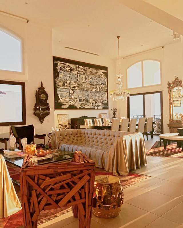 Living room at sunset: a draped tufted sofa, a chair of stacked wood, a gold stool and a bracket clock, the dining room beyond