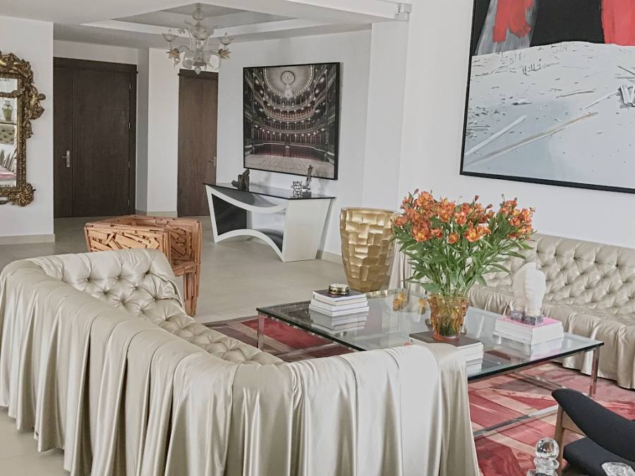 Living room with draped tufted sofas, a glass table with orange lilies, a gold vase and a photograph of a theatre over a white console