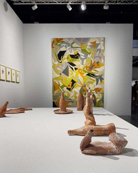 A large felt work in yellow, green and black on the back wall of an art fair booth, clay bottles on a plinth in front