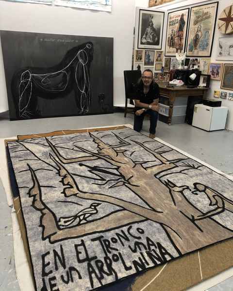 José Bedia kneeling in his studio behind the finished felt En el tronco de un árbol, his paintings on the walls