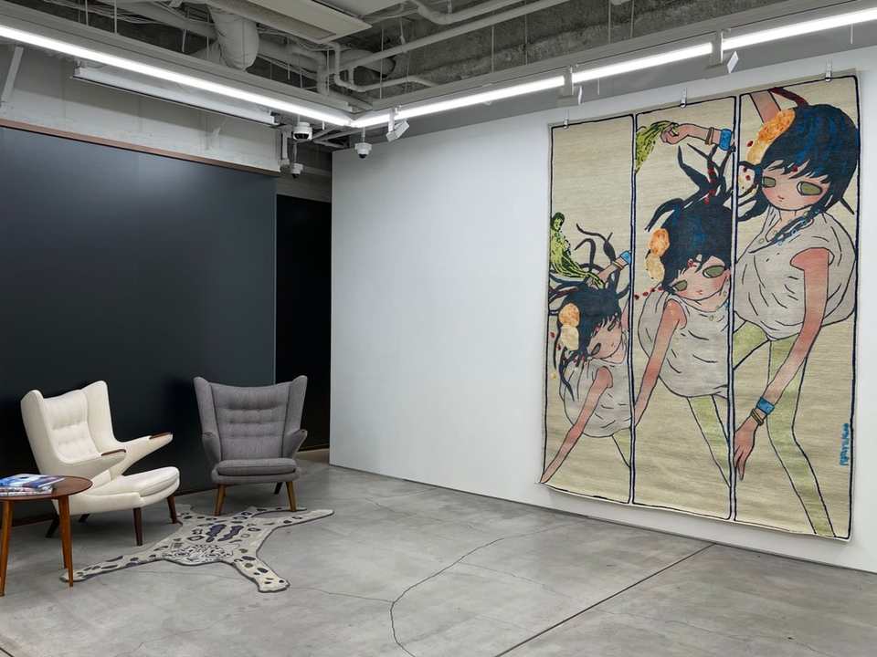 A large hand-knotted work of three girls hanging in a gallery, a leopard-shaped rug on the floor before two armchairs