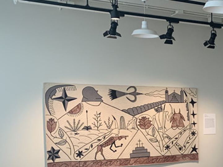 A felt work by José Bedia with a figure, stars, plants and a pyramid in black on cream, hanging in a gallery