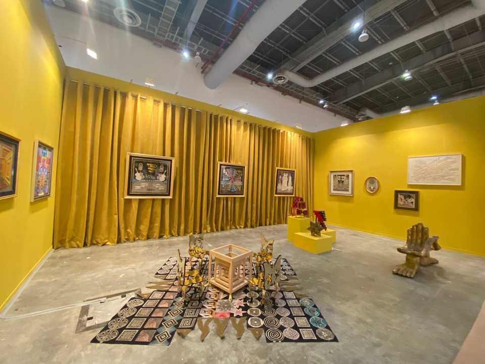 A yellow art fair booth with Pedro Friedeberg's hand-knotted work of symbols on the floor under gilded chairs, his framed works on the walls and the hand chair