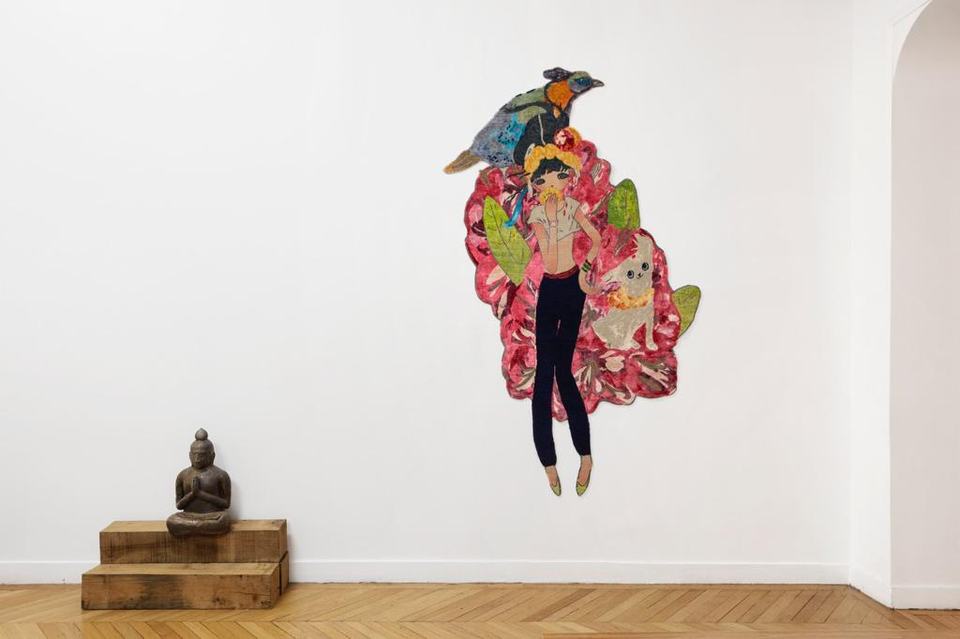 A shaped hand-knotted work by Aya Takano of a girl, a bird and a cat on pink flowers, on a white wall beside a seated stone figure