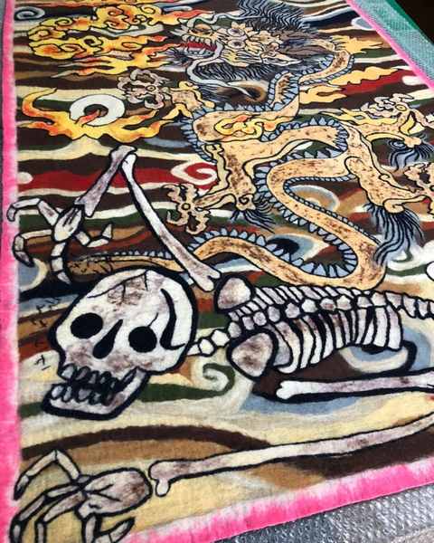 A felt work of a golden dragon in clouds above a skeleton, with a pink border