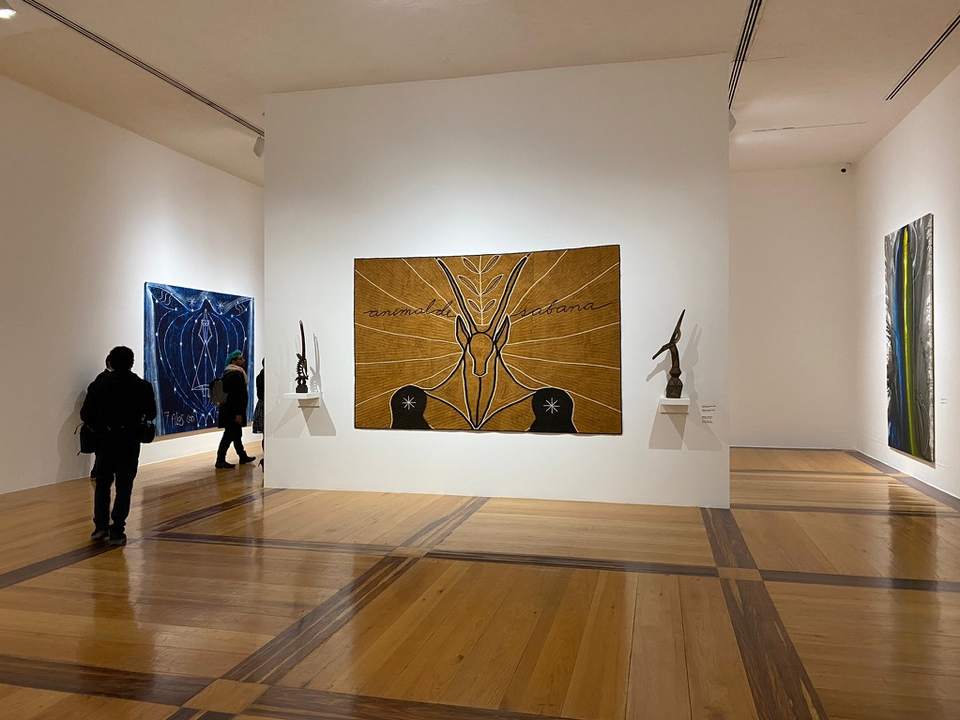 A museum gallery with a large ochre textile of an antelope titled animal di sabana between two sculptures, a blue work on the left