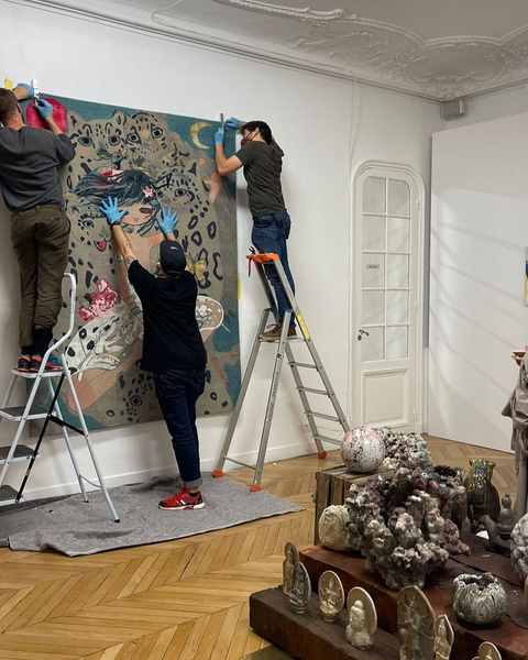Three installers on ladders hanging Aya Takano’s hand-knotted work with leopards in a gallery with a moulded ceiling