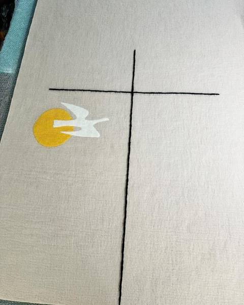 A pale felt panel with a black line cross, a yellow sun and a white dove