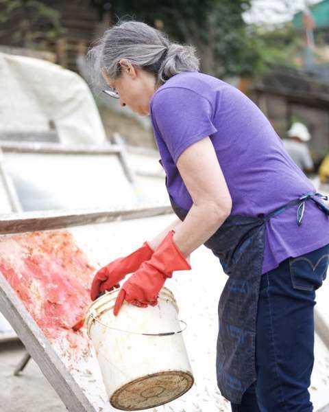 Jessica Stockholder in red gloves pouring red pulp from a bucket onto a tilted mould