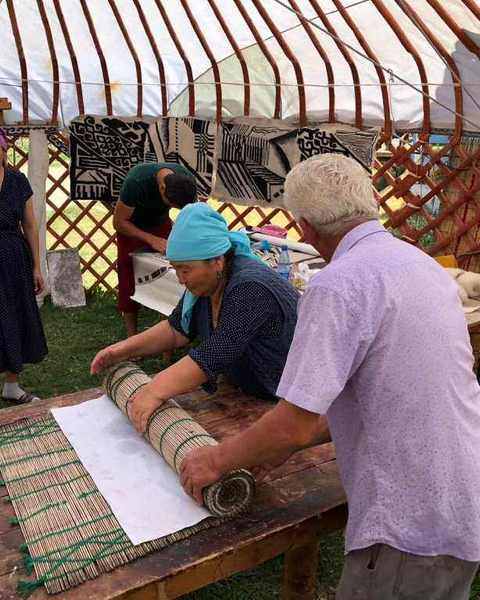 Two felt-makers rolling wool inside a reed mat in a yurt, felt samples hanging behind