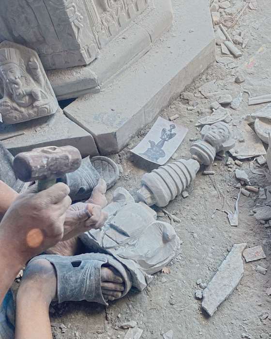 A carver with hammer and chisel seated on the workshop floor, the artist’s printed drawing beside him and the carved column and monkey head on the ground