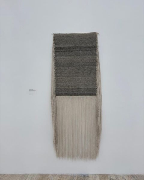 A dark woven panel above a long fall of pale threads hanging on a white museum wall