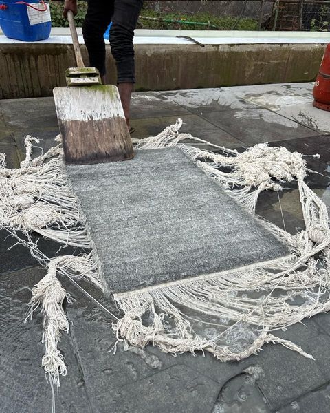 A woven panel with long loose warp threads being washed with a wooden paddle on a wet stone floor