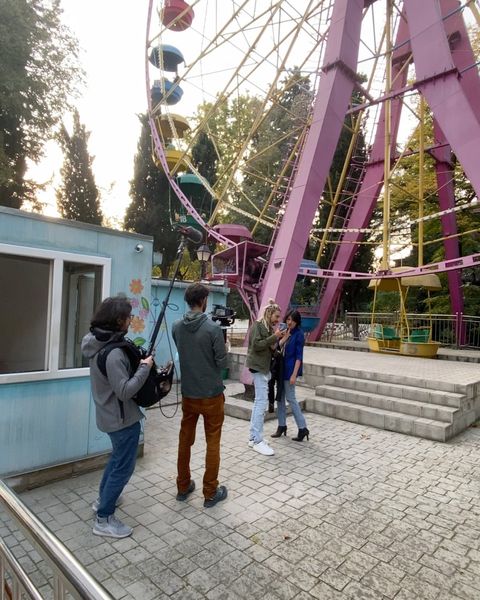 A camera operator and sound recordist filming two women talking beneath a pink Ferris wheel