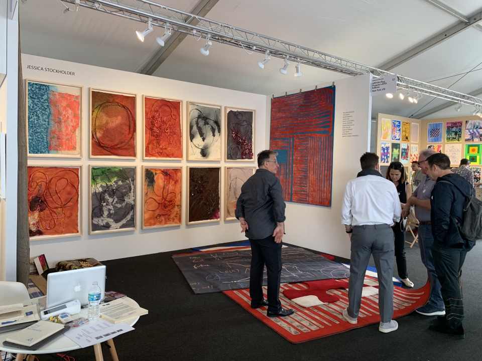 An art fair booth: ten framed lokta paper works by Jessica Stockholder on the back wall, a red and blue hand-knotted work on the right, rugs on the floor