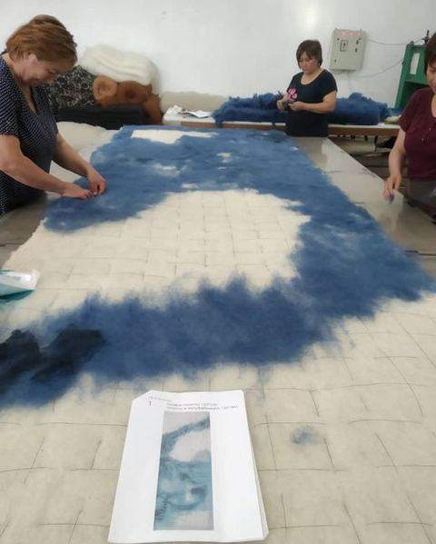 Felt-makers laying blue wool over white on a long table, a printed reference in front