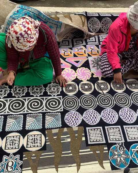 Two women seated on a hand-knotted work of spirals, stars and squares, carving the pile