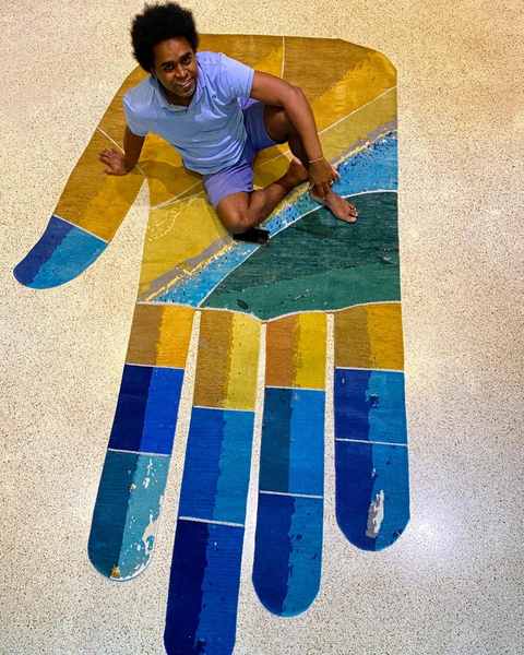A hand-shaped hand-knotted work in yellow, green and blue on a terrazzo floor, a man seated on it