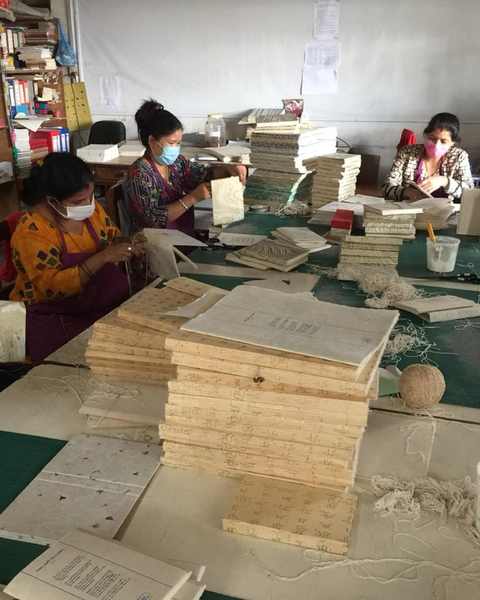 Women in masks binding lokta paper notebooks at long tables
