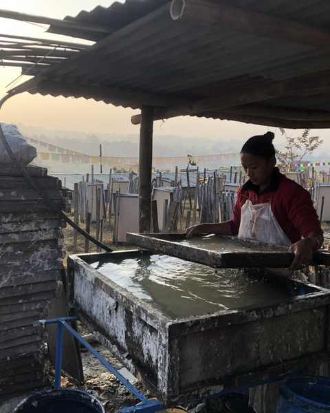 A papermaker lifting a mould from the vat at dawn, rows of drying frames and prayer flags behind