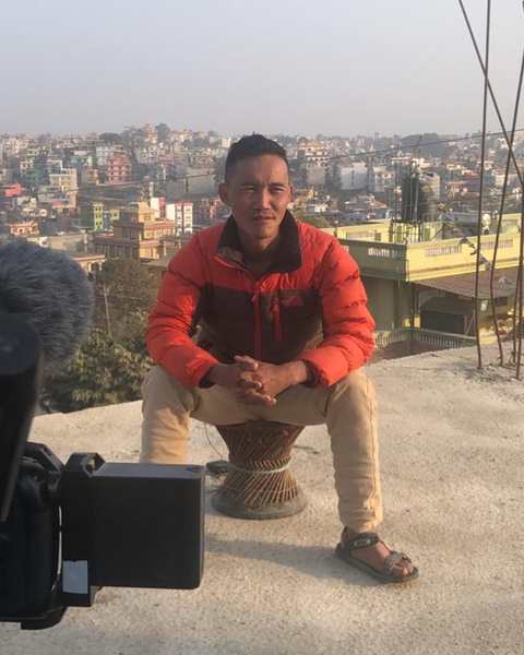 A man in an orange jacket interviewed on a Kathmandu rooftop, camera and boom in the foreground