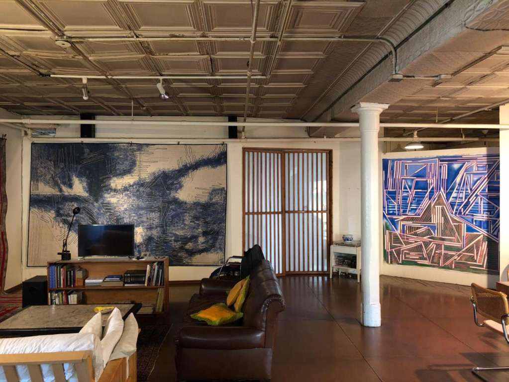 Large textile works hung in a New York loft with a pressed-tin ceiling, a sofa and bookshelves in front