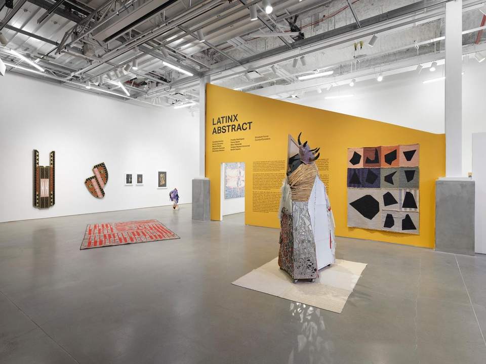 Gallery view of the exhibition Latinx Abstract: a yellow title wall with a felt work of black shapes on pink, blue and grey panels, a sculpture in front, a red rug on the floor