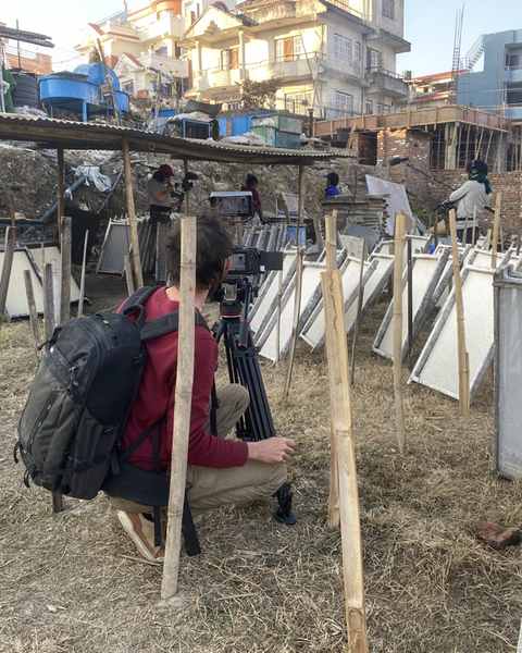 A cameraman crouched behind a tripod among rows of paper moulds drying in a field