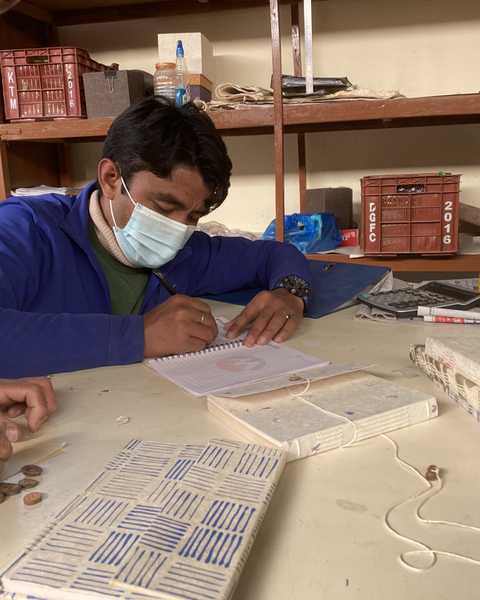 A man in a mask taking notes at a workshop table among lokta notebooks and string