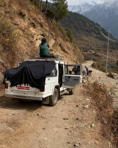A jeep stopped on a dirt mountain road, a sound recordist on the roof, snowy peaks behind