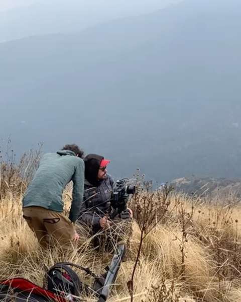 Two crew members filming from a grassy ridge above a misty valley