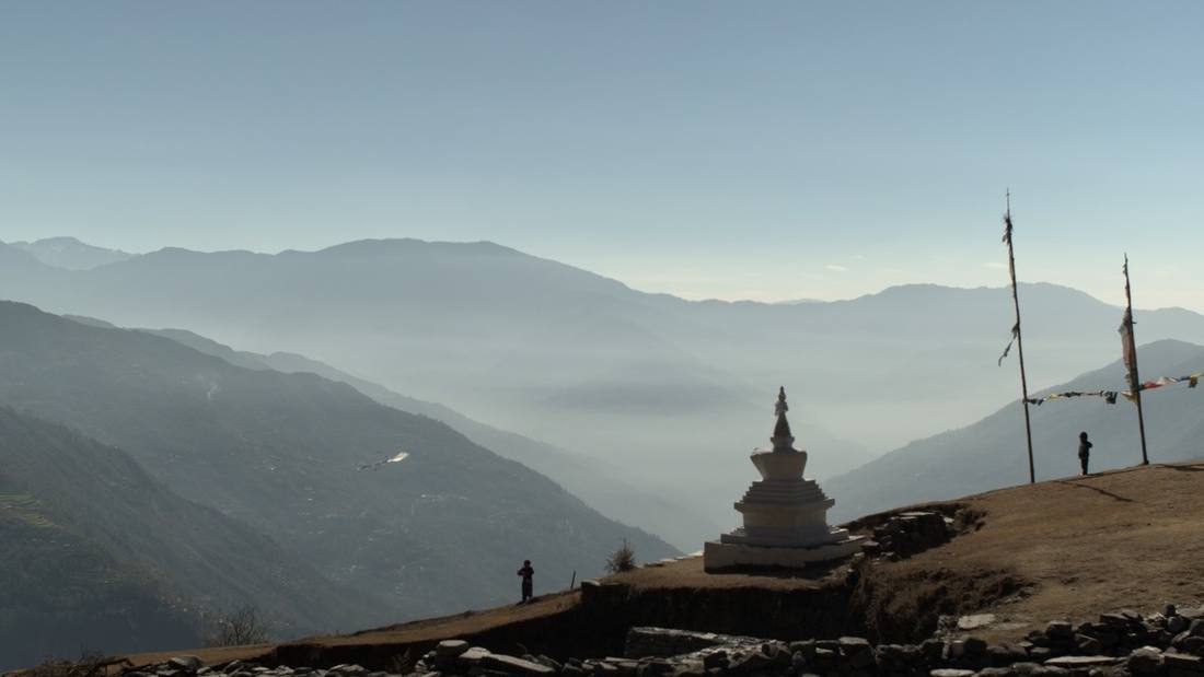 A frame from the film: a white stupa on a hilltop, prayer flags, two small figures and a paper kite over misty Himalayan valleys