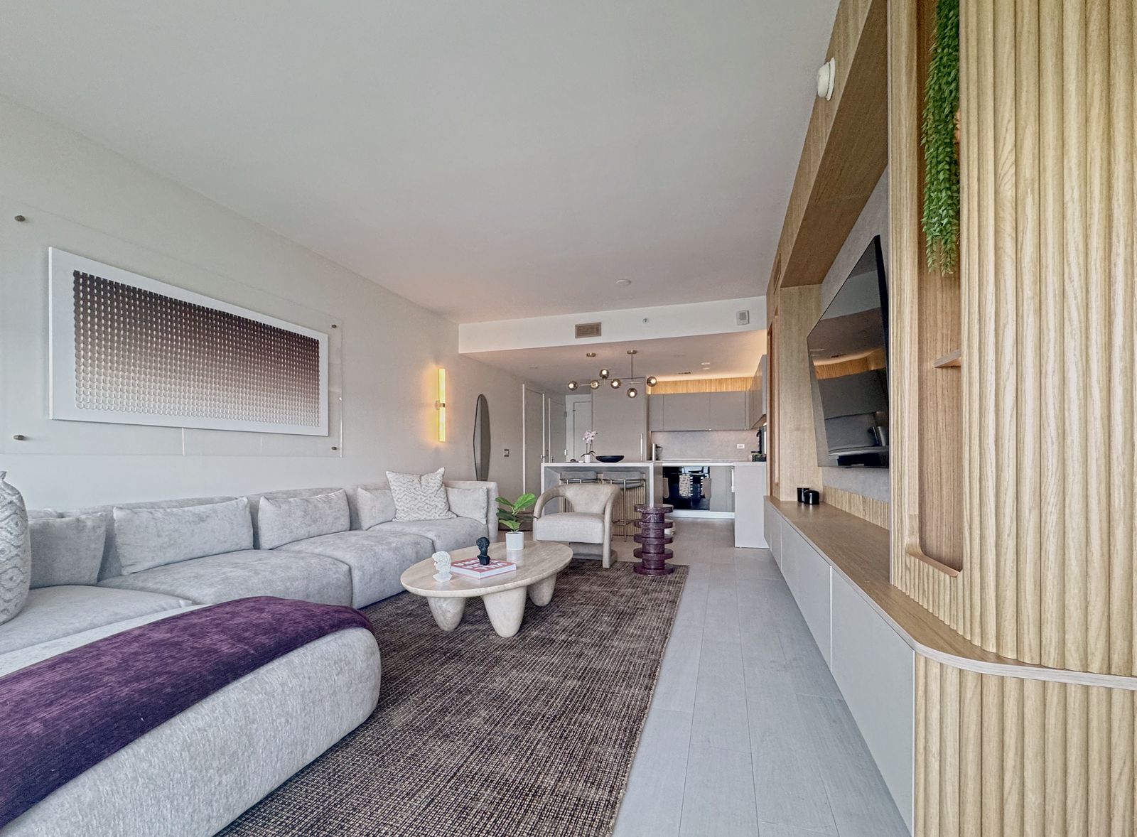 Living room facing the bay: a long pale sectional with a plum throw, a sculptural coffee table and a gradient relief on the wall